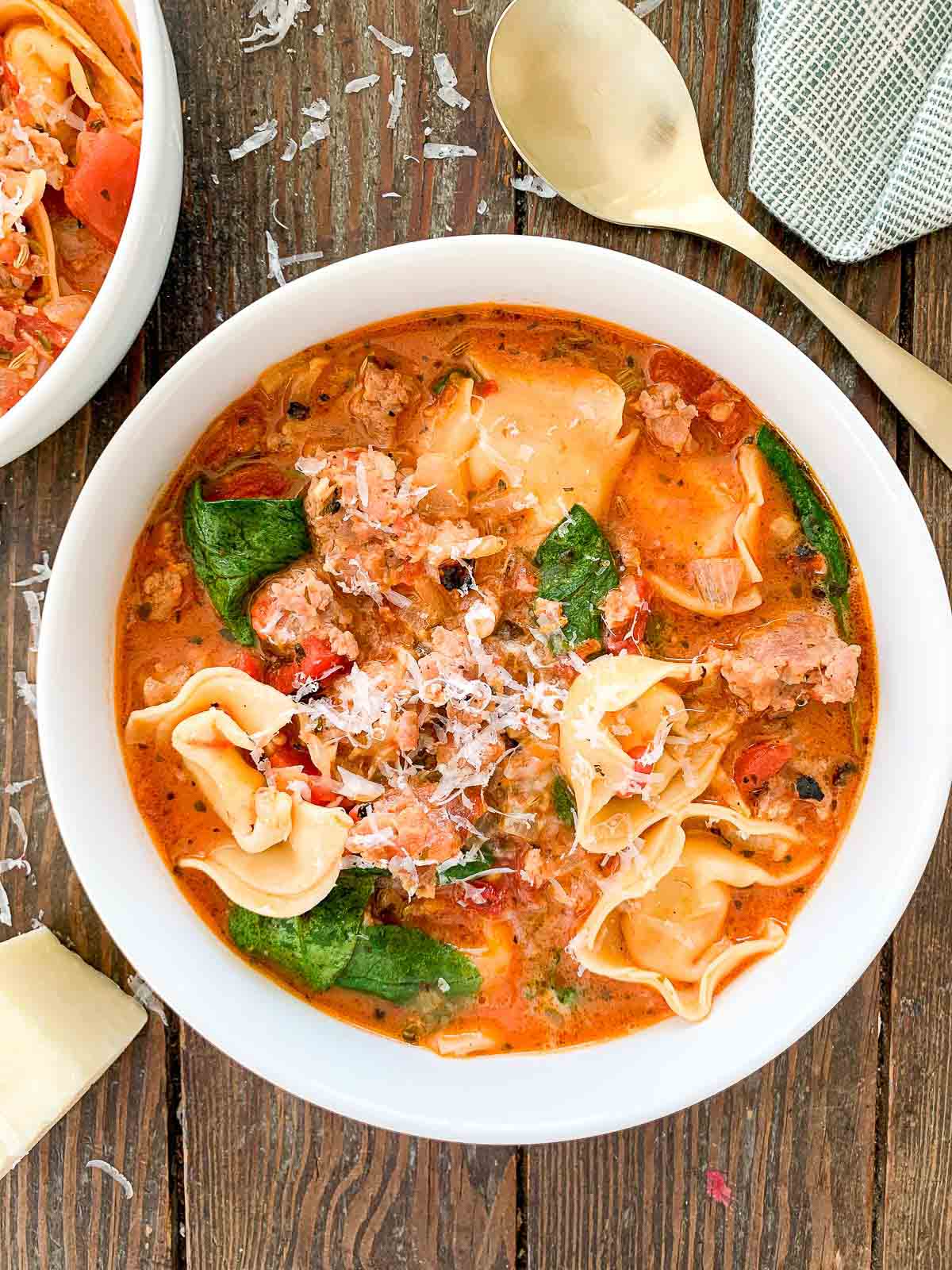 A bowl of creamy tortellini soup with spinach, tomatoes, and ground meat, garnished with grated cheese. A gold spoon and a folded napkin are beside the bowl on a wooden surface.