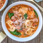 A white bowl filled with creamy tortellini soup, featuring cheese-filled tortellini, ground meat, spinach leaves, and diced tomatoes. The soup is garnished with grated cheese. The bowl sits on a rustic wooden table.