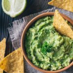 A bowl of creamy guacamole garnished with parsley, accompanied by tortilla chips on a rustic wooden surface. A halved lime is in the background, adding a fresh touch to the scene.