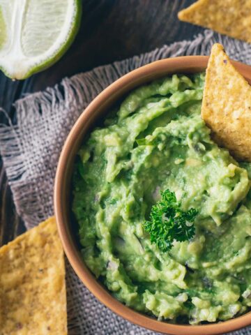 A bowl of creamy guacamole garnished with parsley, accompanied by tortilla chips on a rustic wooden surface. A halved lime is in the background, adding a fresh touch to the scene.