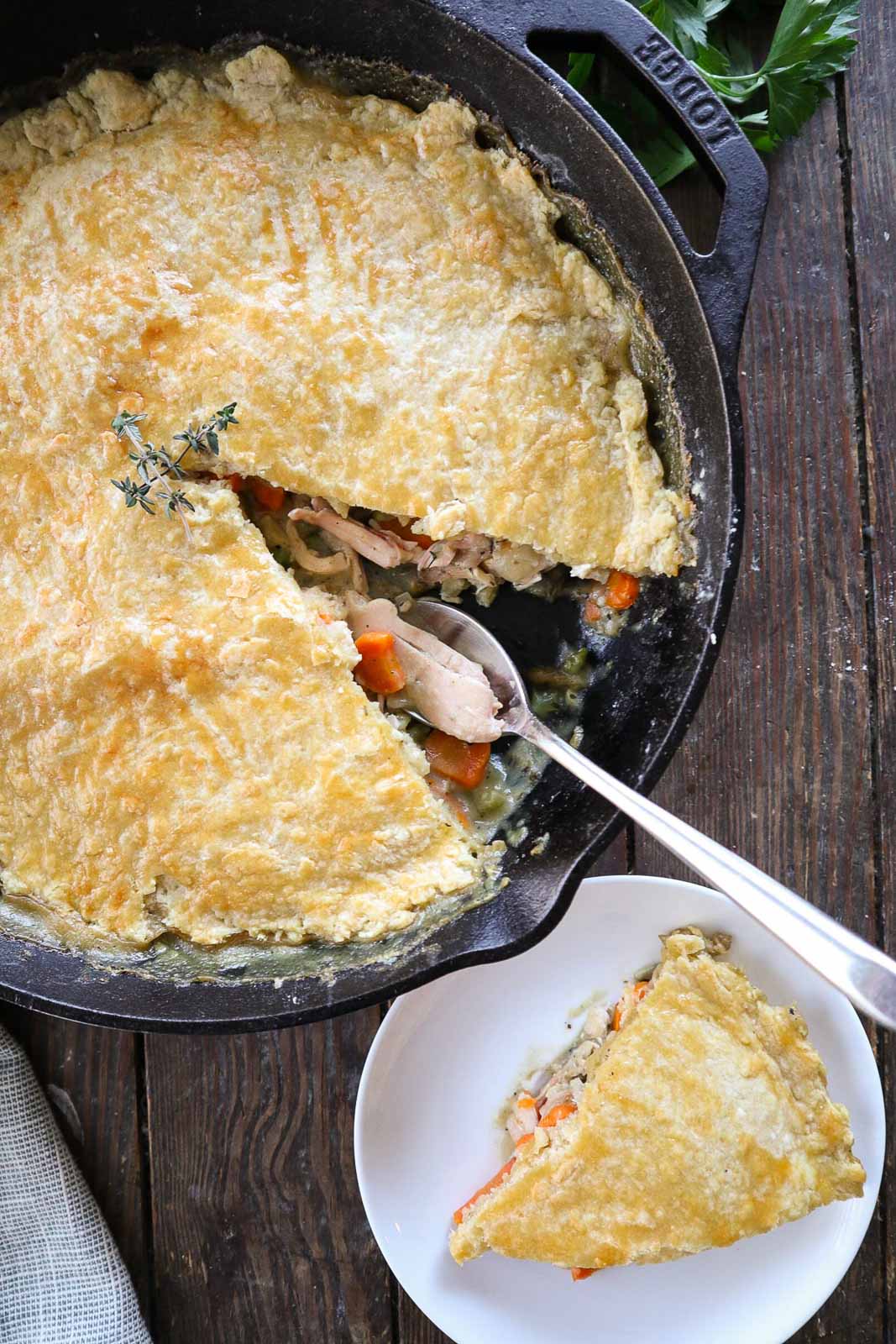A cast iron skillet with a golden-brown sourdough chicken pot pie, partially served. The pie is filled with chicken and vegetables like carrots. A slice has been placed on a small white plate nearby. A sprig of fresh herbs decorates the skillet.