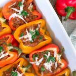 A baking dish filled with red and yellow bell peppers stuffed with a savory meat mixture, topped with melted cheese and garnished with fresh parsley. A whole red bell pepper and a cloth are beside the dish.