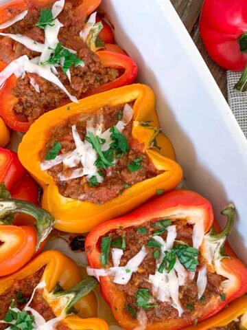 A baking dish filled with red and yellow bell peppers stuffed with a savory meat mixture, topped with melted cheese and garnished with fresh parsley. A whole red bell pepper and a cloth are beside the dish.