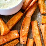Close-up of roasted sweet potato wedges seasoned with herbs and sea salt on parchment paper. A bowl with a creamy dipping sauce is partially visible in the corner. The sweet potatoes have a golden brown color, indicating crispness.