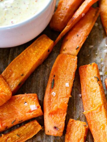 Close-up of roasted sweet potato wedges seasoned with herbs and sea salt on parchment paper. A bowl with a creamy dipping sauce is partially visible in the corner. The sweet potatoes have a golden brown color, indicating crispness.