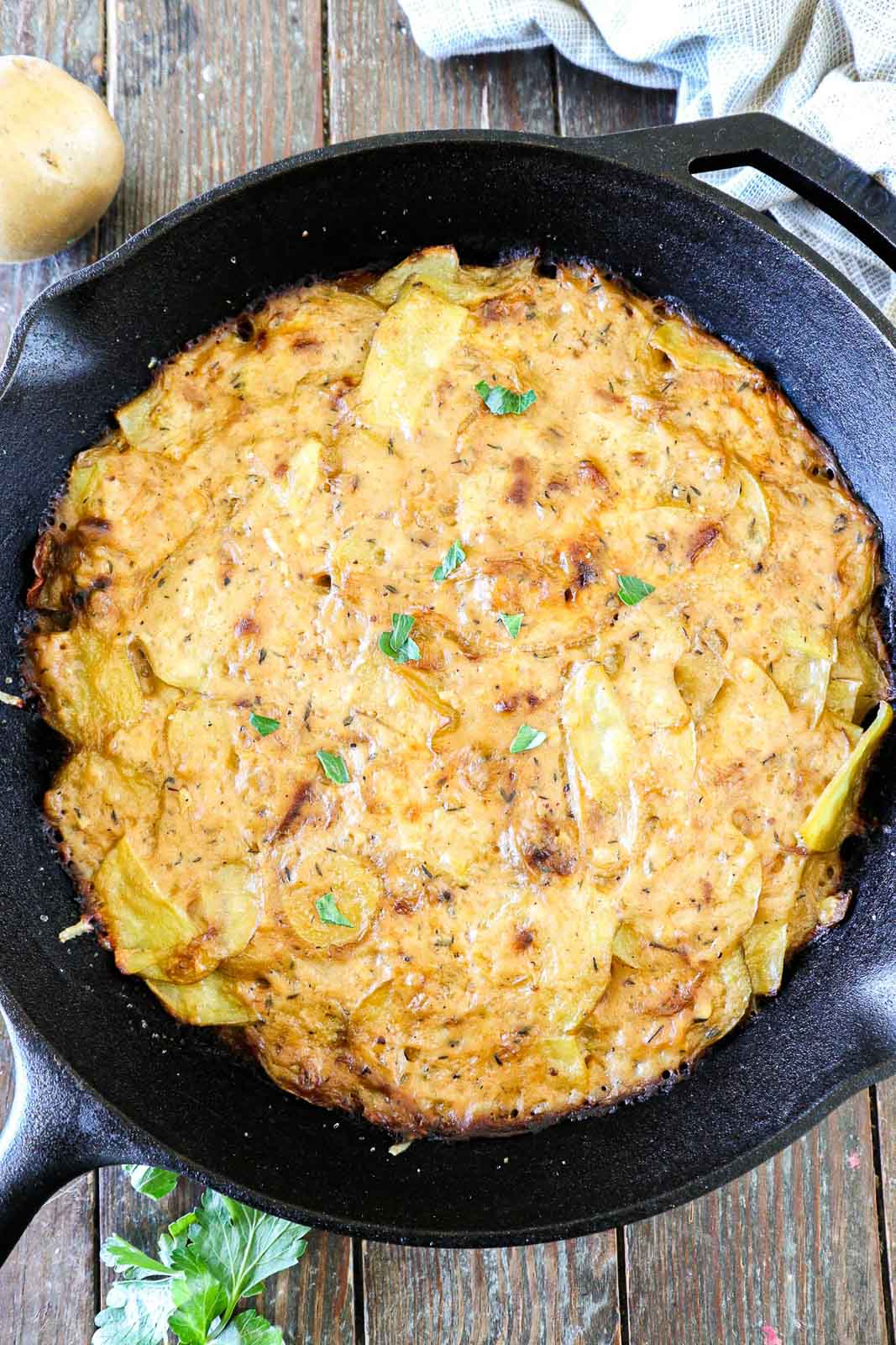 A cast iron skillet filled with baked scalloped potatoes, topped with golden, melted cheese and garnished with fresh parsley. The skillet rests on a wooden surface, with a hint of a cloth and a potato nearby.