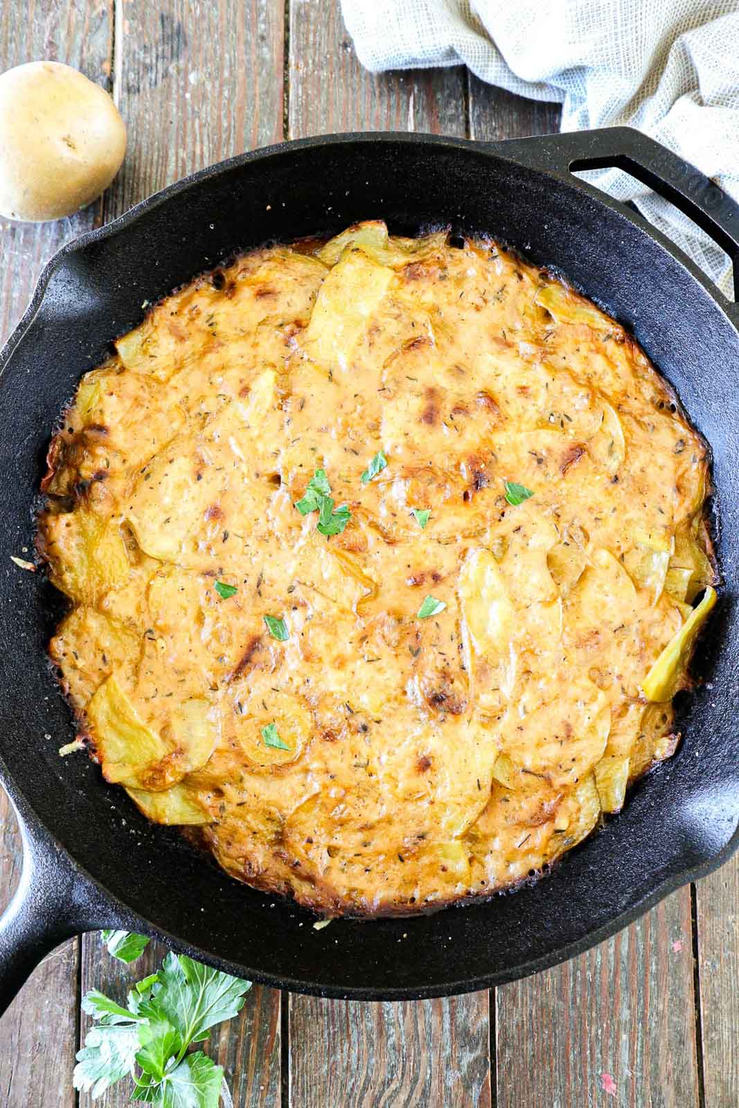 A skillet filled with golden brown scalloped potatoes garnished with parsley. The dish appears creamy and baked, with a crispy top layer, placed on a rustic wooden surface. A potato and a sprig of parsley are nearby.