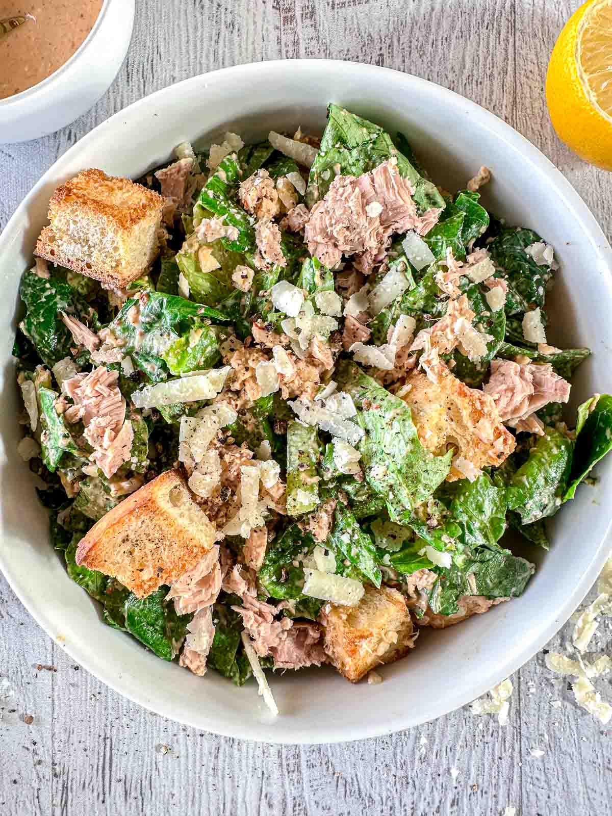 A bowl of Caesar salad with leafy greens, chunks of tuna, croutons, grated cheese, and a sprinkle of black pepper. A small bowl of dressing and a lemon wedge are beside the bowl on a light wooden surface.
