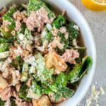 A close-up of Tuna Caesar salad with lettuce, chunks of tuna, croutons, grated cheese, and a dusting of black pepper. A lemon wedge is visible at the top right corner on a light wooden surface.
