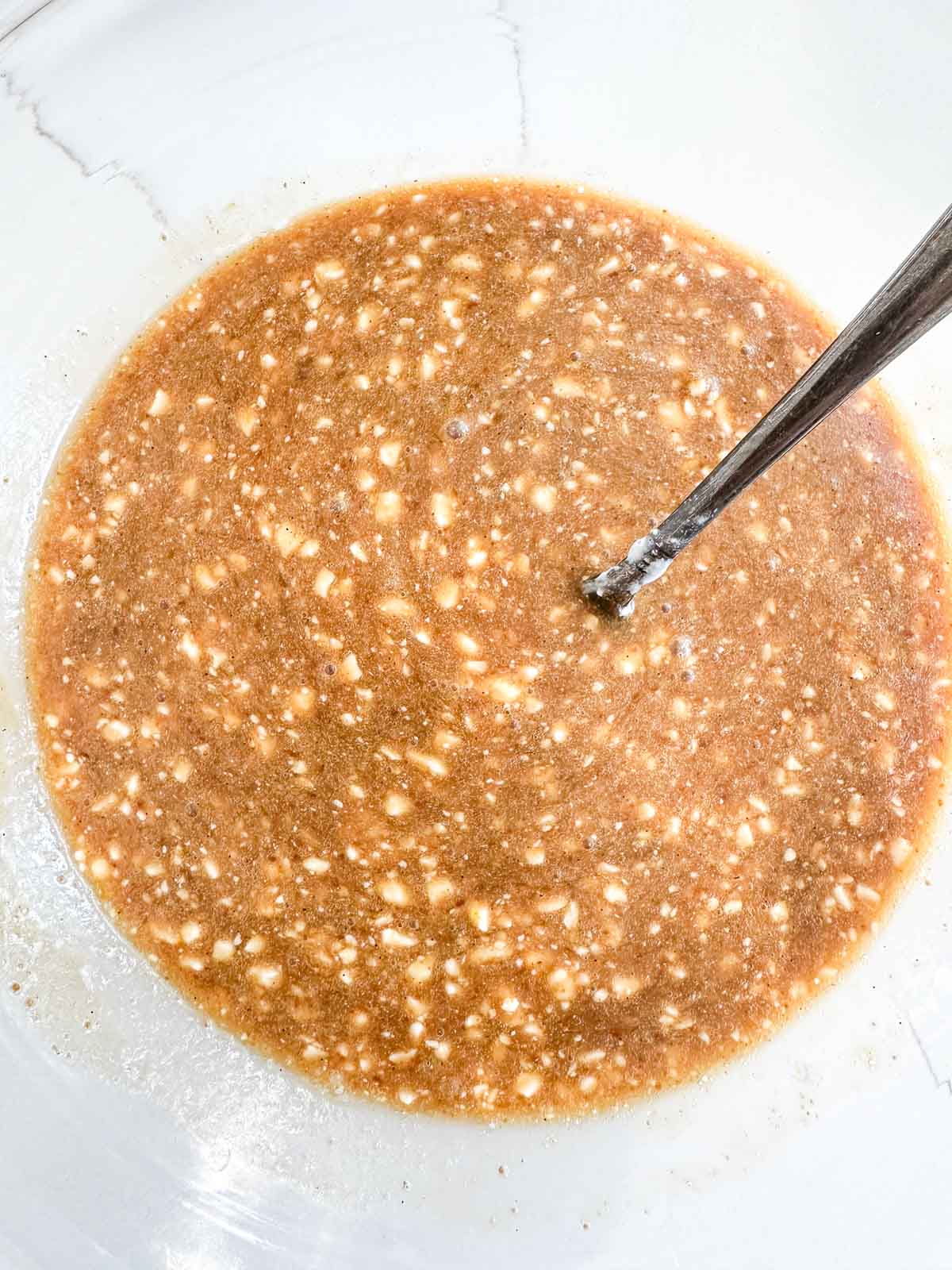 A close-up of a mixing bowl containing the wet ingredients for cottage cheese blueberry muffins. A metal spoon is partially submerged in the mixture.