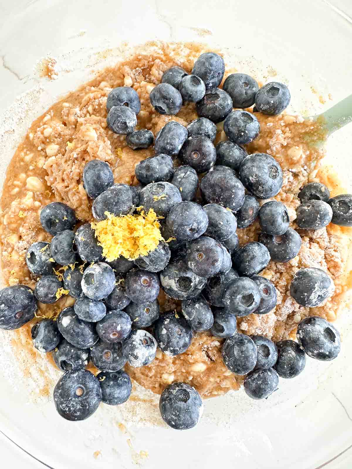 A mixing bowl with batter contains a heap of fresh blueberries and a small mound of lemon zest on top.