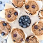 Eight freshly baked cottage cheese blueberry muffins with blueberries are arranged around a small bowl of fresh blueberries. Each muffin is wrapped in brown parchment paper.