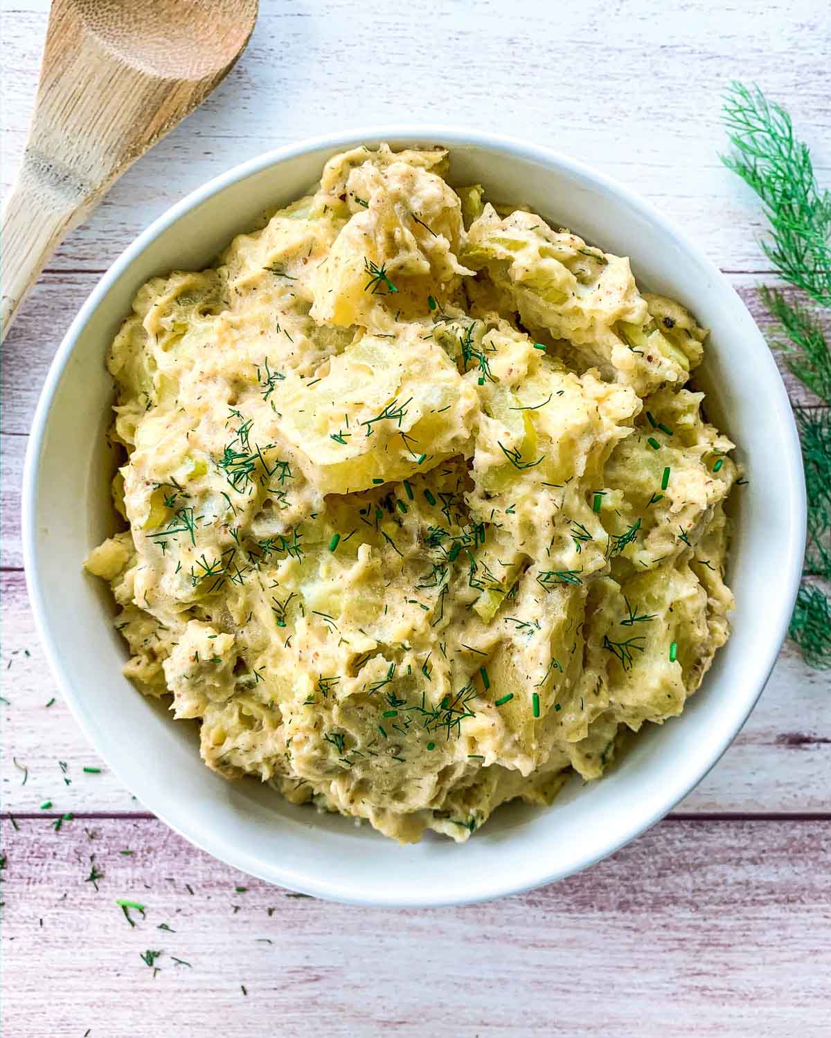 A bowl of creamy potato salad garnished with fresh dill. The salad is light yellow, featuring chunks of potatoes and herbs. A wooden spoon and sprigs of dill are nearby on a light wooden surface.