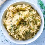 A bowl of creamy potato salad garnished with fresh chopped dill. The salad is chunky and appears to have a seasoning blend. Its set on a light speckled surface with a sprig of dill nearby.