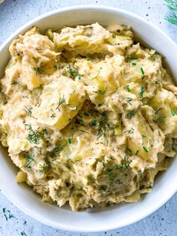 A bowl of creamy potato salad garnished with fresh chopped dill. The salad is chunky and appears to have a seasoning blend. Its set on a light speckled surface with a sprig of dill nearby.