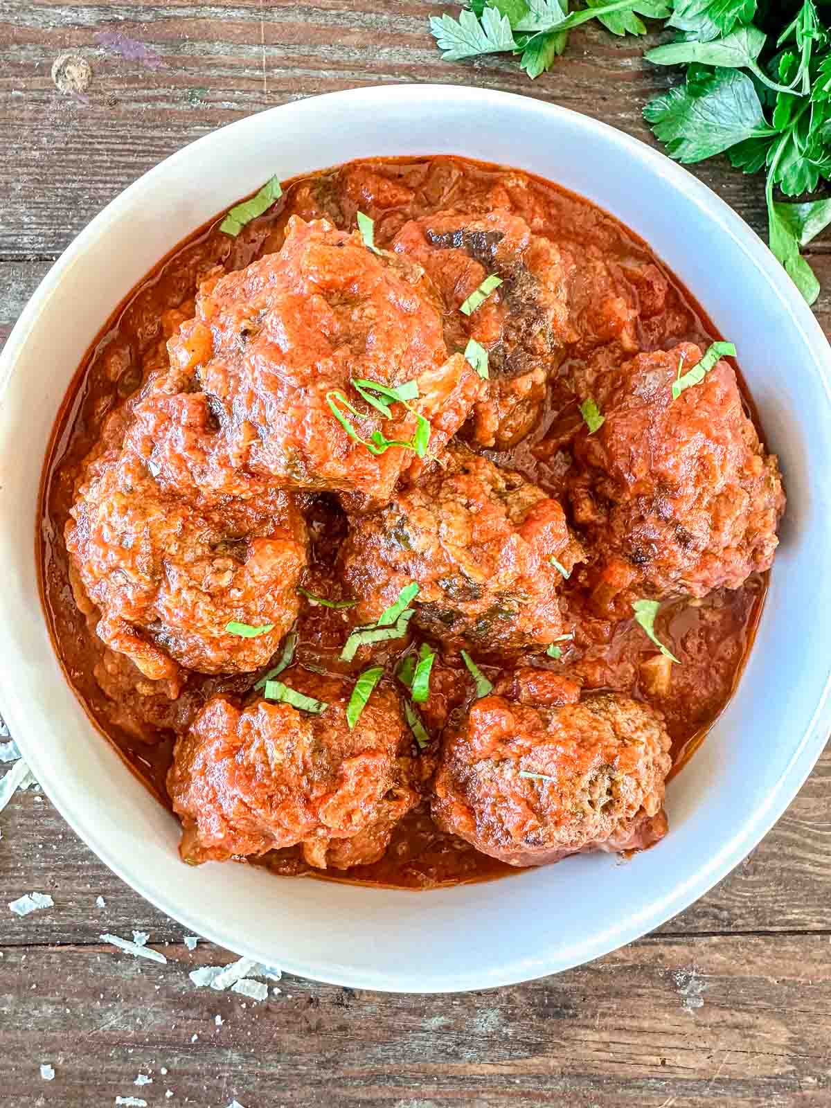 A white bowl filled with meatballs in a rich tomato sauce, garnished with chopped fresh herbs. The bowl is placed on a wooden surface, with sprigs of parsley nearby.