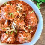 A bowl of meatballs covered in tomato sauce, topped with grated cheese and garnished with fresh basil.