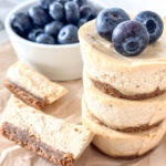 Stack of creamy mini cheesecakes topped with blueberries, placed on parchment paper. A bowl of blueberries is in the background. One cheesecake slice is positioned on its side, displaying its layers.