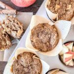 Three apple sourdough muffins in paper liners sit in a baking tray next to chopped apple pieces in another compartment. A sliced muffin with apple chunks is on a plate nearby, alongside two cinnamon sticks and a whole apple.