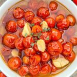 Close-up of a bowl filled with glistening cooked cherry tomatoes, garlic cloves, and sprigs of fresh thyme. The vibrant red and glossy texture of the tomatoes is highlighted, creating an appetizing and flavorful appearance.