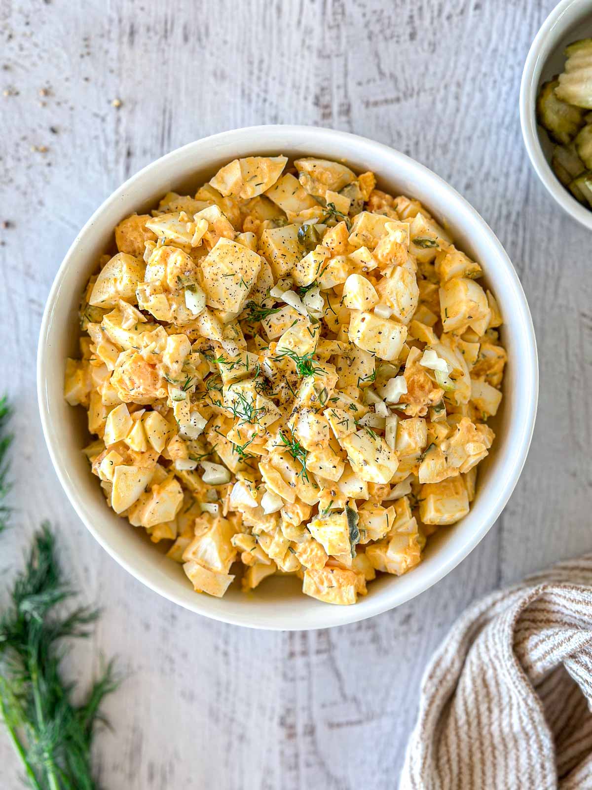 A bowl of creamy potato salad garnished with fresh dill and black pepper is on a light wooden surface. Some herbs and a striped cloth are partially visible nearby, adding to the rustic presentation.