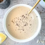 A creamy gluten free caesar dressing in a white bowl sprinkled with black pepper, on a light wooden surface. A gold spoon is dipping into the sauce. A lemon wedge and a bowl partially filled with green salad are nearby.