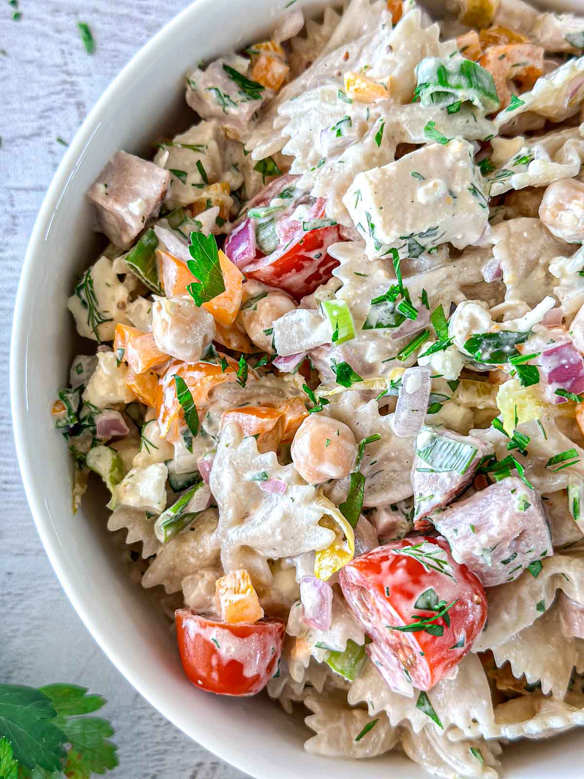 A bowl of creamy pasta salad with bowtie pasta, cherry tomatoes, chickpeas, diced ham, bell peppers, red onion, and fresh herbs. The salad is garnished with chopped parsley and dill. The bowl is placed on a light-colored surface.