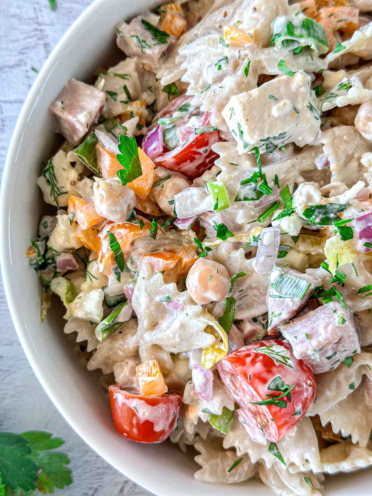 A close-up of a bowl of creamy pasta salad. It features farfalle pasta, cherry tomatoes, chickpeas, diced peppers, and fresh herbs, all coated in a creamy dressing. Fresh parsley is sprinkled on top for garnish.