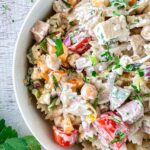Close-up of a creamy high protein pasta salad in a white bowl, featuring farfalle pasta, diced tomatoes, bell peppers, red onion, parsley, and chickpeas. The salad is garnished with fresh herbs and appears lightly dressed in a creamy sauce.