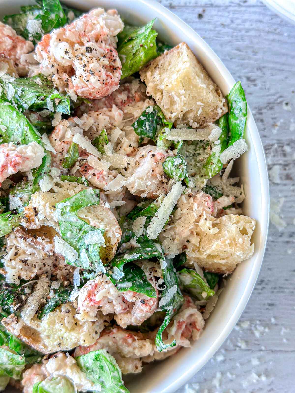Close-up of a Caesar salad in a white bowl, featuring romaine lettuce, croutons, shredded Parmesan cheese, and a pepper seasoning. A creamy dressing lightly coats the ingredients. The bowl rests on a textured grey surface.