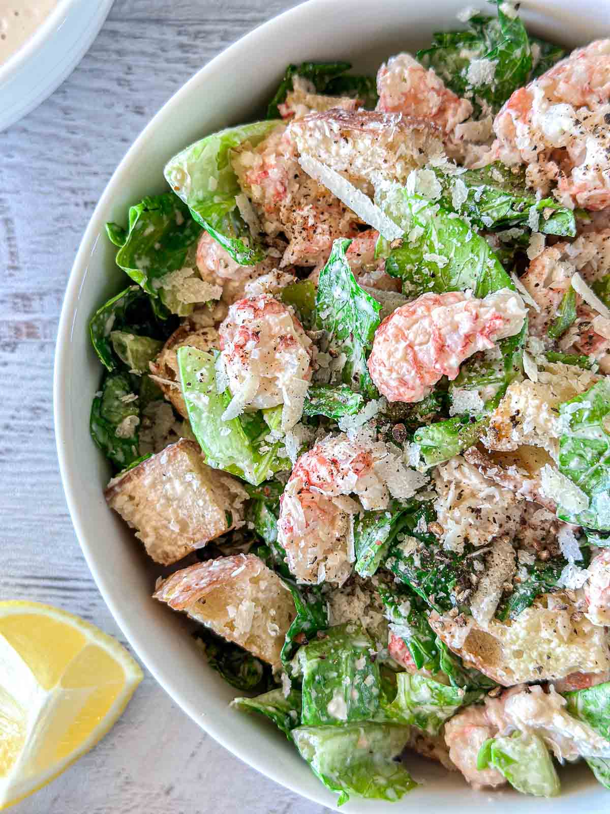 A bowl of Caesar salad with chunks of lobster, romaine lettuce, croutons, and grated parmesan cheese. The salad is garnished with ground pepper and accompanied by a lemon wedge on the side.