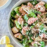A bowl of Lobster Caesar salad with lettuce, croutons, lobster pieces, and grated cheese, topped with ground pepper.