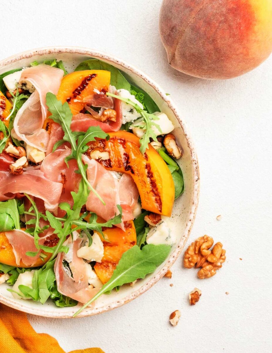 Grilled peach and burrata salad with prosciutto on a white background.
