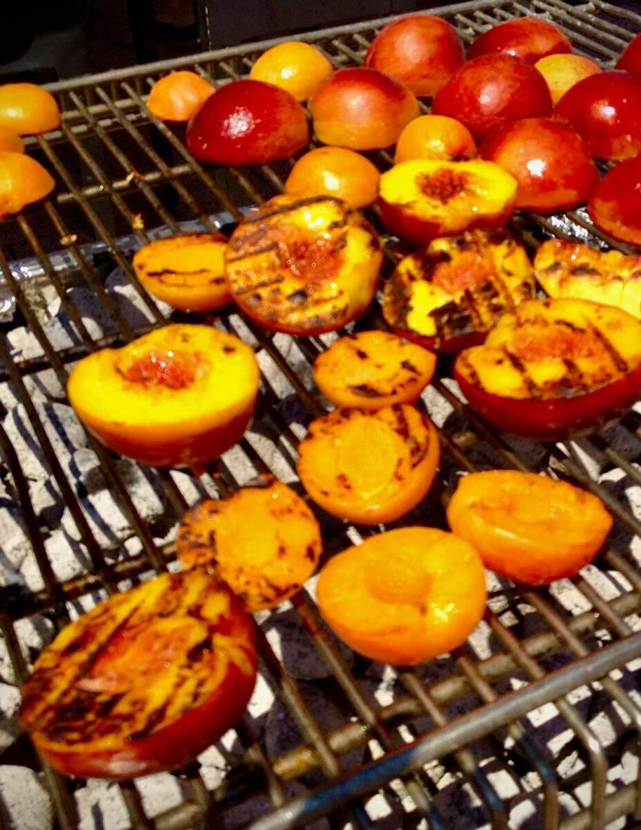 Halved peaches are being grilled on a barbecue. The fruits have char marks and are amidst glowing charcoal beneath the metal grill.
