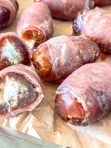 Close-up of prosciutto-wrapped dates with some cut open to reveal a creamy filling. The dates are placed on parchment paper, alongside part of a Wüsthof Classic knife.