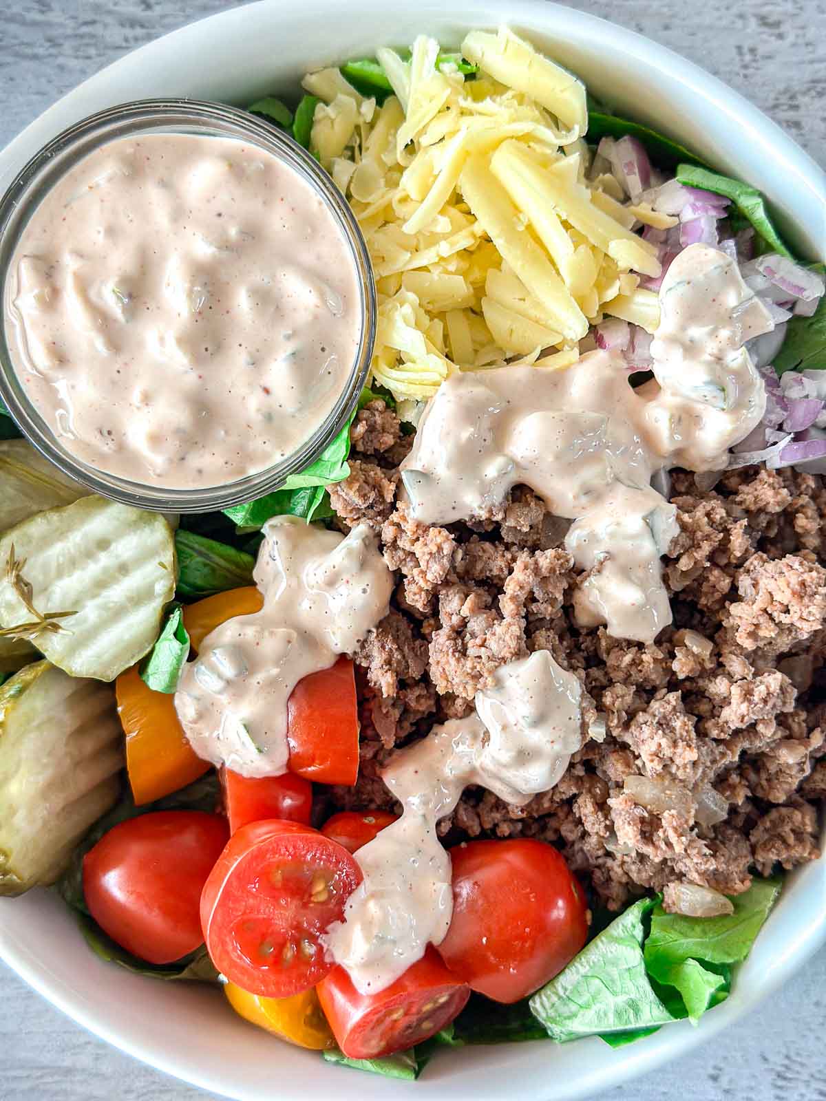 A bowl of salad containing seasoned ground beef, cherry tomatoes, pickles, shredded cheddar cheese, diced onions, and chopped lettuce. A small dish of creamy dressing is placed on the side, with additional dressing drizzled on the salad.