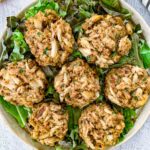 A plate of six baked Dungeness crab cakes, garnished with chopped herbs, is arranged on a bed of leafy greens.