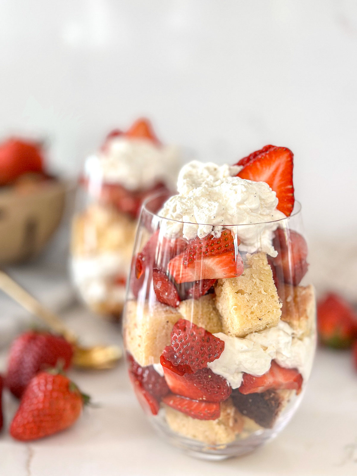 Two clear glasses filled with layers of cubed pound cake, sliced strawberries, and whipped cream, garnished with a fresh strawberry on top. A basket of strawberries and a gold spoon are in the background on a white surface.
