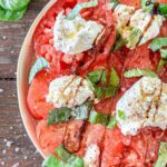 Sliced tomatoes topped with dollops of creamy burrata, fresh basil leaves, cracked black pepper, and a drizzle of balsamic glaze, arranged on a plate on a rustic wooden surface.