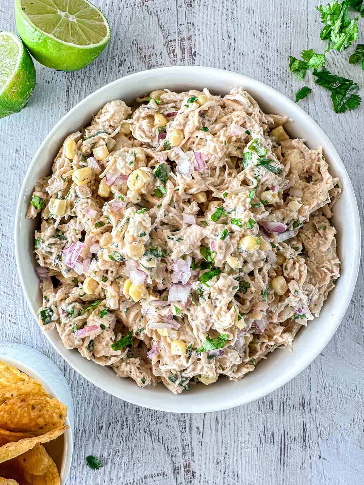 A bowl of creamy chicken salad with corn, red onion, and fresh herbs, placed on a light wooden surface. Lime halves, tortilla chips, and chopped herbs are nearby.