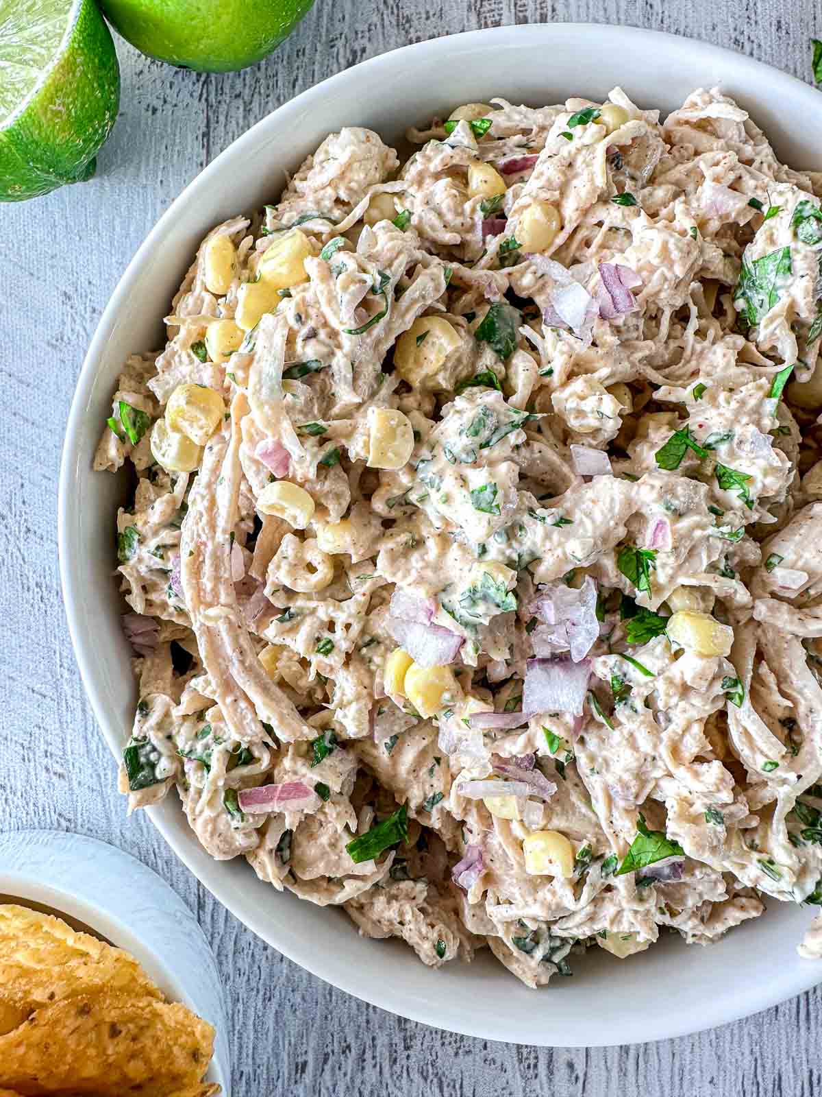 A white bowl filled with creamy shredded chicken salad mixed with corn, red onion, cilantro, and herbs, placed on a light wooden surface. Lime halves and tortilla chips are nearby.