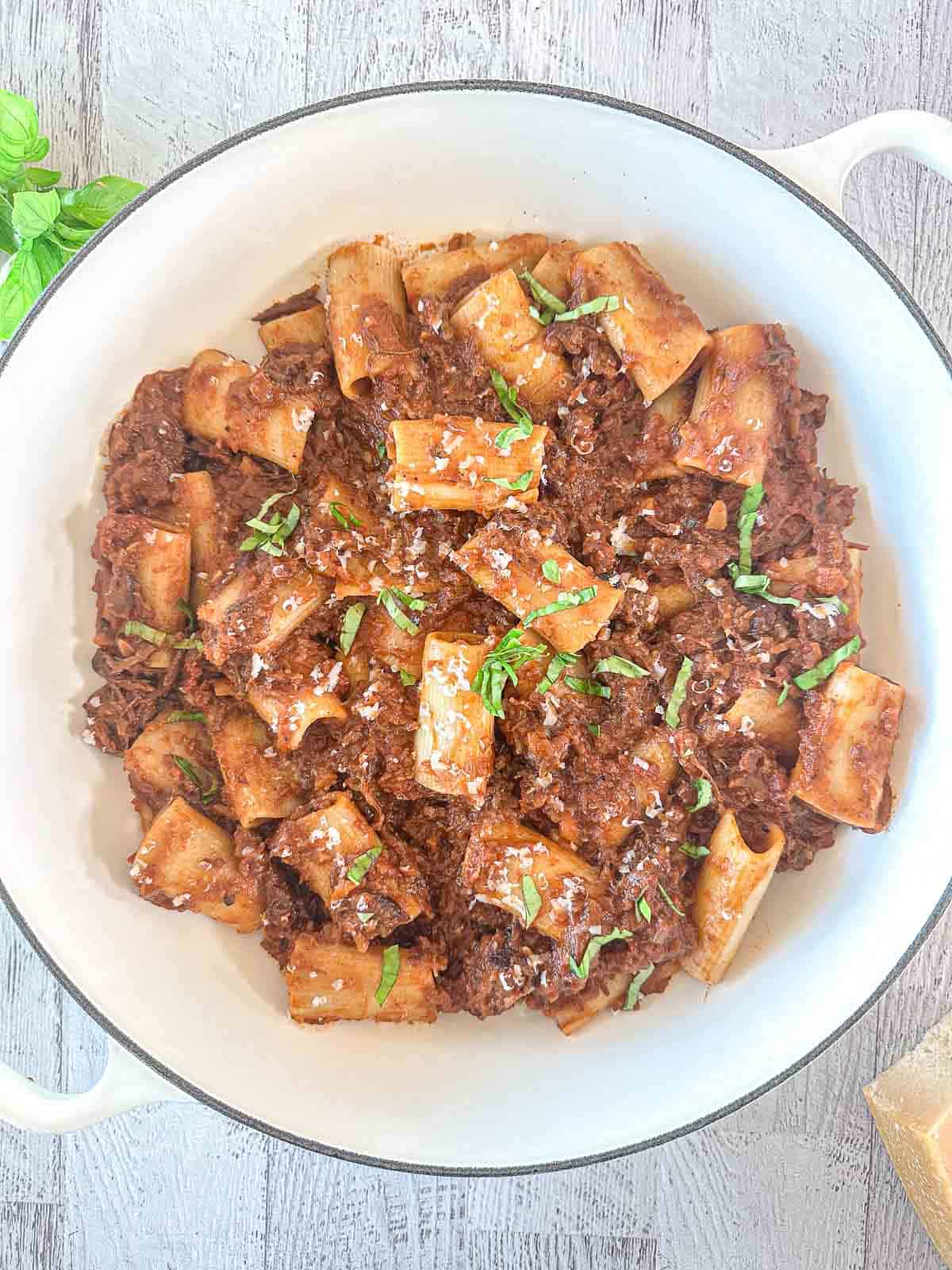 A white bowl filled with rigatoni pasta topped with a rich, meaty red sauce, garnished with grated cheese and chopped fresh herbs, sits on a light wooden surface.