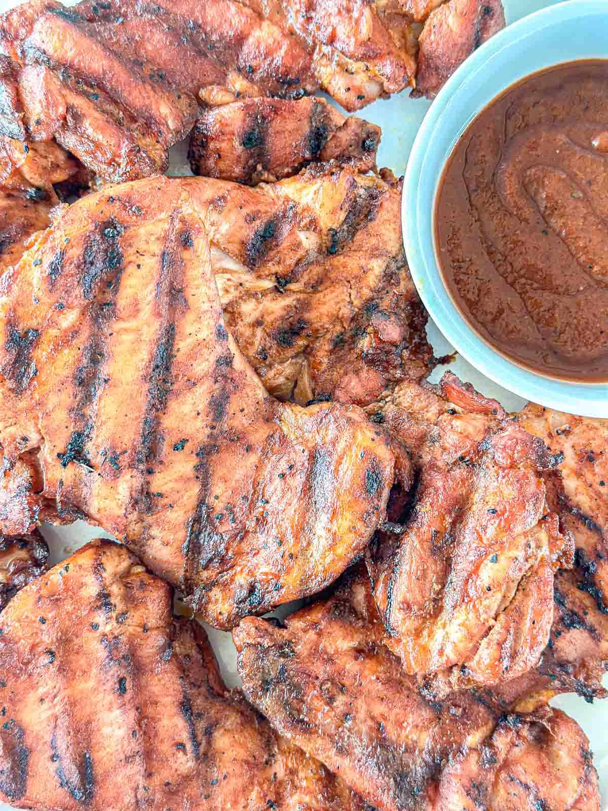Grilled, marinated chicken breasts with dark grill marks are arranged on a surface next to a bowl of brown barbecue sauce. The chicken looks juicy and well-seasoned.