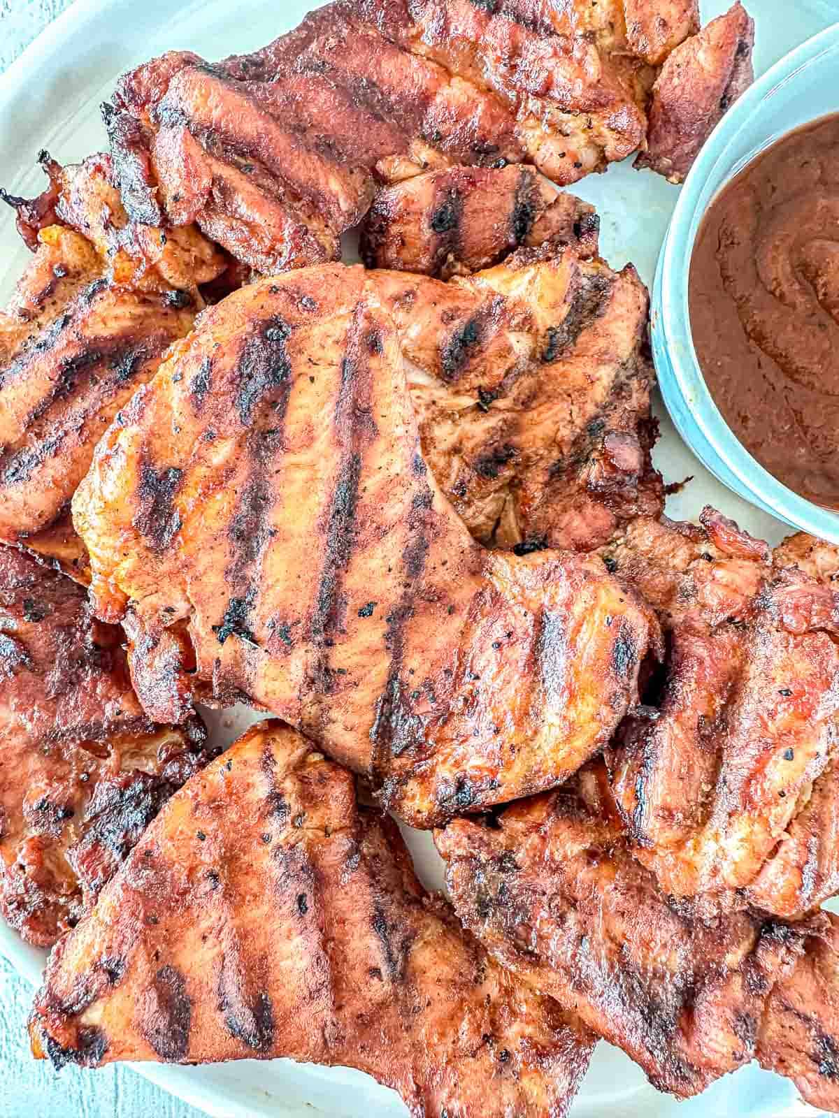 A plate of grilled, marinated chicken pieces with visible char marks, served next to a small bowl of reddish-brown dipping sauce.