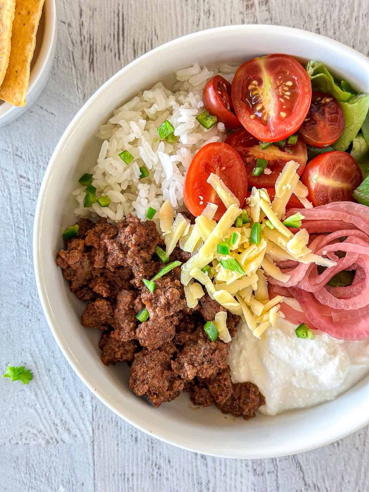 A white bowl filled with ground beef, white rice, shredded cheese, halved cherry tomatoes, pickled onions, chopped green onions, lettuce, and a dollop of sour cream, arranged in separate sections.