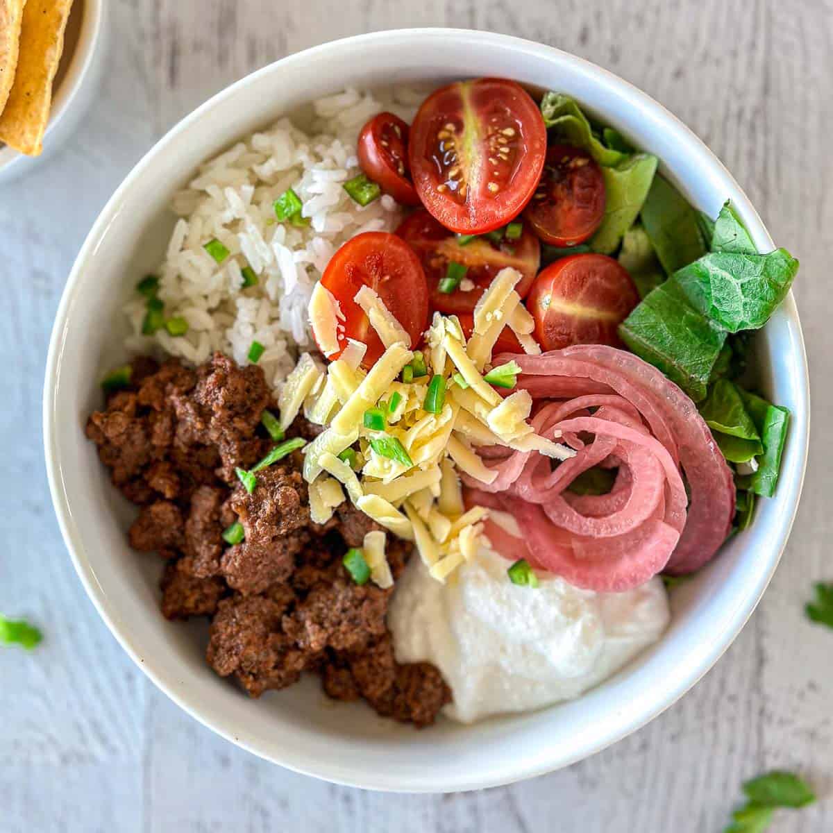 A cottage cheese taco bowl with white rice, ground beef, shredded cheese, halved cherry tomatoes, spinach, pickled red onions, and a dollop of cottage cheese, garnished with chopped green onions on a light wooden surface.