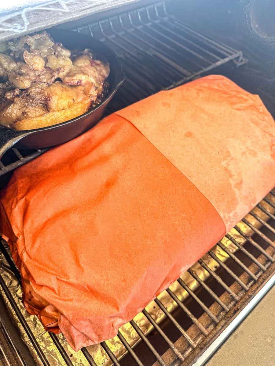 Beef Brisket wrapped in butcher paper on the grill grates of a Traeger grill. A small cast iron of beef tallow is next to it.