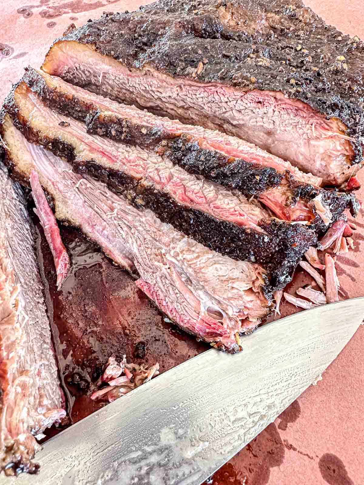 Sliced smoked brisket with a dark, peppery bark and juicy, pink interior sits on a cutting board next to a large knife, with juices visible around the meat.