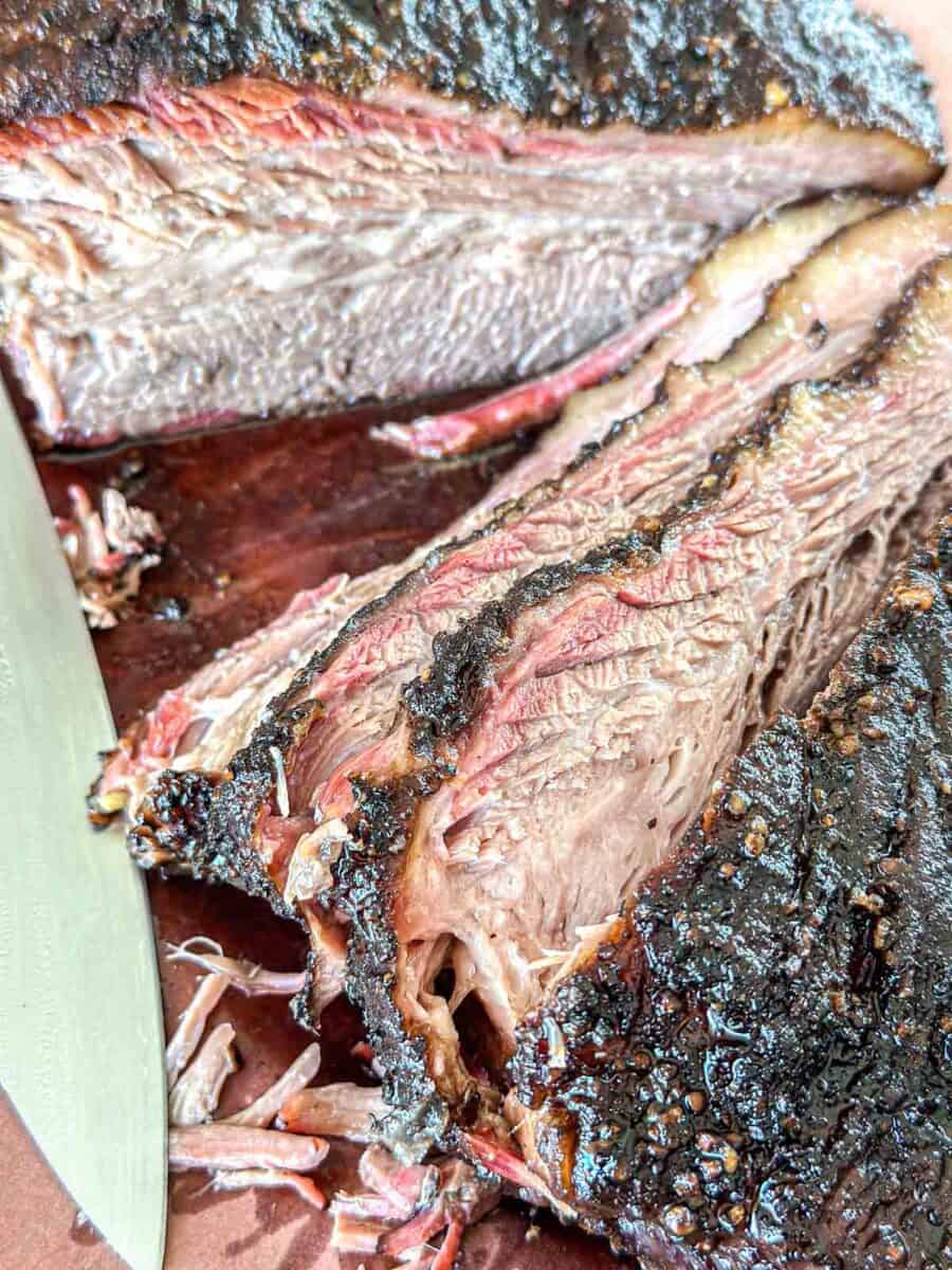 Close-up of sliced smoked brisket on a cutting board, showing a dark, seasoned crust, pink smoke ring, and juicy, tender meat with a knife partially visible beside the slices.