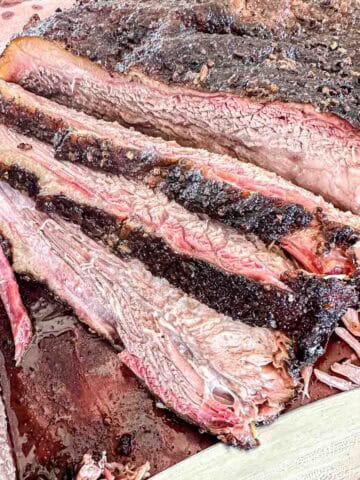 Close-up of sliced smoked brisket showing a dark, seasoned crust and juicy, tender interior with visible smoke ring layers on a cutting board.